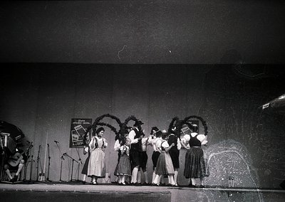 Five women in traditional Bavarian attire perform on stage, holding decorative wreaths. Stage lighting and microphones sugges...