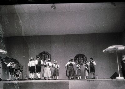 Group in traditional Alpine attire performing on stage, likely 1950s–1970s. Men wear vests, hats, and aprons; women in dirndl...