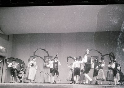 Mid-20th century folk dance performance in traditional Alpine attire. Stage setup includes floral arches, a microphone stand,...