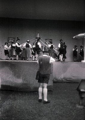 Indoor folk dance performance featuring traditional attire (dirndls, vests, hats) and musicians playing accordions. Stage set...