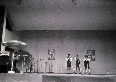 Four male performers in matching suits and hats play instruments on a stage with microphones, microfiche posters, and a drum ...