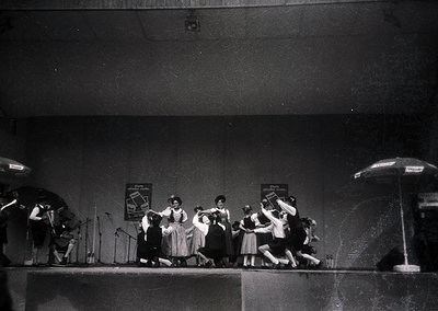 Mid-20th century stage performance featuring a group dance with musicians playing brass instruments. Women in knee-length dre...