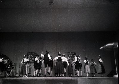 Black-and-white stage performance featuring traditional folk dancers in coordinated attire—skirts, blouses, and headscarves—w...