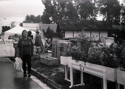Black-and-white street scene featuring a woman in a striped sweater carrying bags, walking past a temporary market stall with...