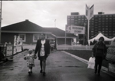 Black-and-white street scene featuring a woman in a striped coat and hat holding a child’s hand, walking toward a "Rodinné Do...
