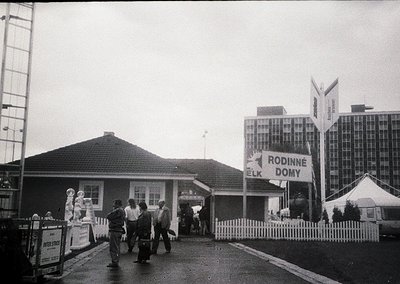 Black-and-white shot of a 1960s-era Czech family resort entrance, featuring a small wooden building with a peaked roof and a ...