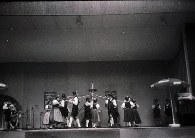 A black-and-white stage performance featuring a group of dancers in traditional folk attire, likely Eastern European, perform...