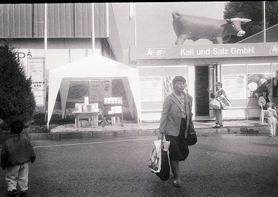 Mid-20th century street scene outside a **Kali und Salz GmbH** storefront, likely Germany. A woman in a structured suit carri...