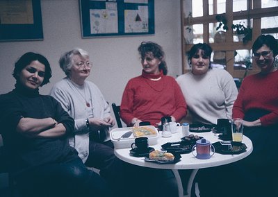 Four women seated around a round table in an indoor café, likely from the **1980s–1990s**. Warm lighting, retro decor with fr...