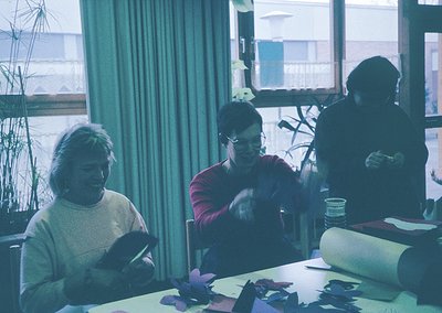 Vintage group crafting session indoors, likely 1970s–1980s. Three women seated at a table cutting paper shapes, surrounded by...