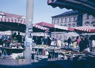 Vibrant 1960s-70s outdoor market with striped red-and-white canopies, showcasing fresh produce and flowers. Historic European...