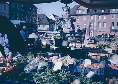 Vibrant 1970s European outdoor market with fresh produce—root vegetables, greens, and crates—displayed under blue plastic cov...