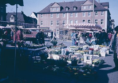 Vibrant 1960s European street market with flower stalls under striped awnings, set against a brick building with dormer windo...