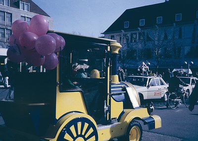 Vintage yellow balloon vendor cart with pink cluster balloons, parked on a European street. Mid-20th century urban scene feat...