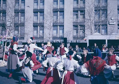 Traditional folk dancers in vibrant, embroidered costumes perform in an urban square, likely Bulgaria, 1960s-70s. Men wear re...