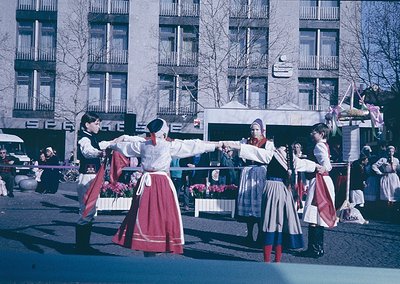 Traditional folk dancers in coordinated red skirts, white blouses, and embroidered vests perform outdoors in an urban setting...