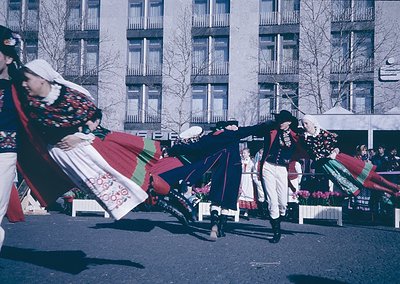 Traditional folk dancers in vibrant, embroidered costumes perform outdoors in a Soviet-era urban setting. Men in dark vests, ...