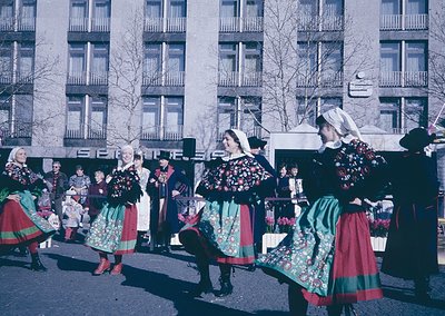 Traditional folk dancers in vibrant, embroidered costumes perform in an urban setting, likely Eastern Europe, 1960s-70s. Gree...