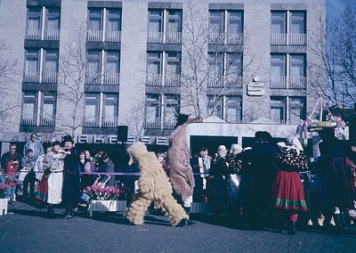 Vintage street scene featuring traditional folk performers in Eastern European attire, likely Bulgaria. A masked dancer in a ...