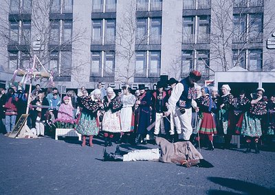 Traditional folk dance performance in urban setting, likely Bulgaria. Group in colorful embroidered costumes with men in whit...
