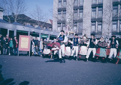 Traditional folk dancers in colorful, embroidered costumes perform in an urban square, likely during a cultural festival. The...