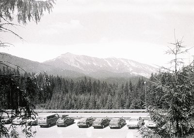 Mid-20th century mountain roadside parking area with classic cars, likely 1970s–1980s. Snow-covered pavement and evergreen fo...