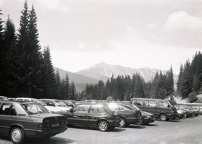 Black-and-white shot of a mountain parking lot with 1980s-era sedans and station wagons, likely European. Snow-capped peaks a...
