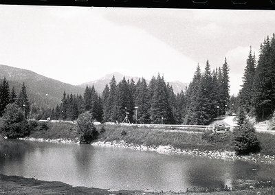 Mid-20th century black-and-white shot of a winding road alongside a serene alpine lake, framed by dense coniferous forests an...