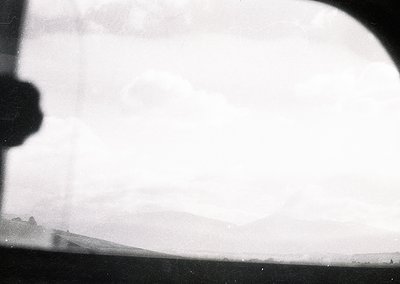 Vintage aerial view through a curved aircraft window, showing hazy terrain below. Distinctive low-lying clouds obscure detail...