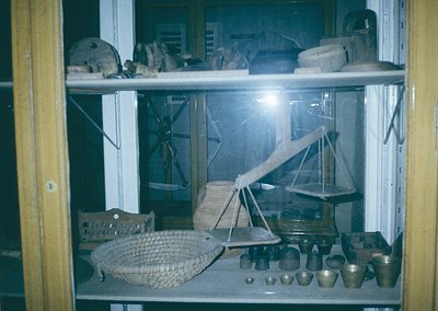 Vintage glass display case showcasing traditional handcrafted tools and pottery. Visible items include a large woven basket, ...