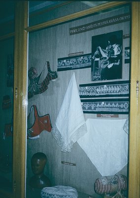 Vintage folk costume display featuring embroidered textiles, traditional footwear, and headwear. Text in Czech suggests cultu...