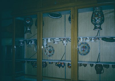 Vintage ceramic display showcasing ornate plates, pitchers, and teapots with floral/geometric patterns. Glass-front cabinet s...