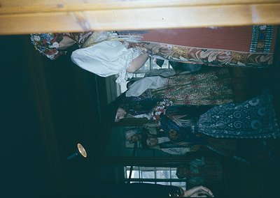 Vintage train compartment with two passengers seated in bunk beds. Upper bunk features floral-patterned fabric and a white pi...