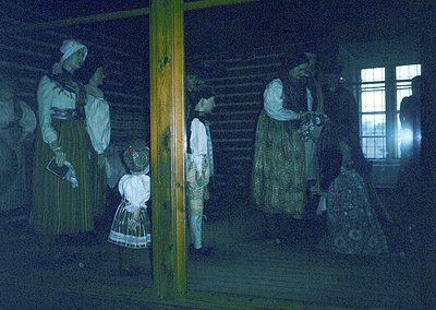 Mannequins in traditional folk attire inside a rustic wooden interior, likely a museum exhibit. Dark, earthy tones dominate w...