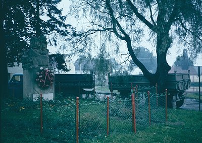 Vintage cemetery scene with Soviet-era monument featuring hammer-and-sickle emblem, surrounded by red-painted metal posts. Ov...