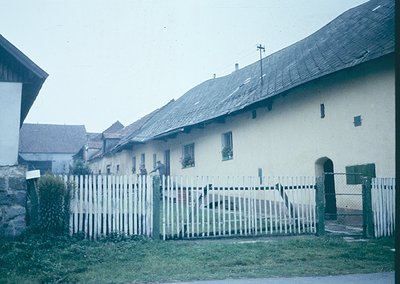 Vintage rural house with whitewashed walls and dark slate roofs, framed by a picket fence. Mid-20th century European farmstea...