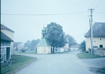 Vintage rural street scene featuring a small Orthodox chapel with a green dome and white walls, flanked by traditional wooden...
