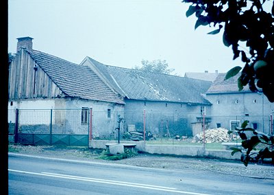 Vintage rural house with weathered brick and tiled roof, surrounded by chain-link fencing. Overgrown vegetation and construct...
