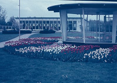 Vintage 1960s-70s camping resort entrance featuring geometric tulip beds in red, white, and blue. Modernist pavilion with gla...