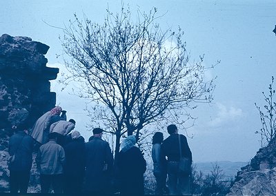 Group of 12+ individuals in winter attire (scarves, coats) gathered on rocky terrain, overlooking a distant landscape. Leafle...
