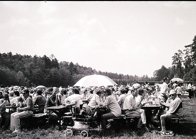 Black-and-white outdoor gathering in a wooded area, likely mid-20th century. Crowded picnic scene with groups seated on bench...
