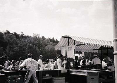 Black-and-white shot of a bustling outdoor food stall, likely from the **1950s–1960s**. Striped canopy with "MARSHMALLOW" sig...