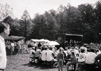 Outdoor concert or event in a wooded park setting, featuring a stage truck and seated audience on wooden benches. Mid-20th ce...
