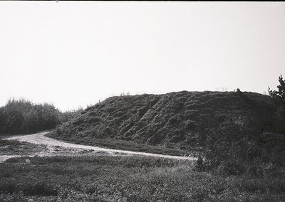 Dense, eroded earthen mound with layered, stepped terrain alongside a narrow, unpaved road. Vegetation includes sparse shrubs...