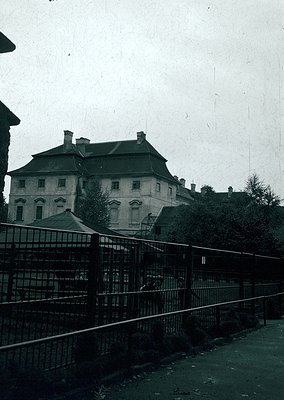 Neoclassical building with symmetrical façade, arched windows, and slate roof behind wrought-iron fence. Likely institutional...