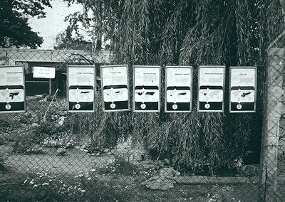 Black-and-white posters depicting firearms with Cyrillic text, mounted on a chain-link fence. Likely Soviet-era propaganda or...
