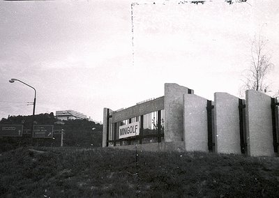 Vintage black-and-white photo of a concrete barrier with "MINGOLF" sign, likely a 1960s-70s roadside advertisement. Urban lan...