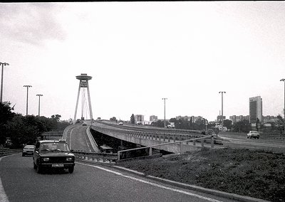 Vintage black-and-white shot of a futuristic bridge tower rising over a highway in an urban setting. Mid-20th century vehicle...