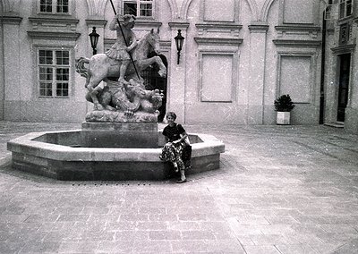 Neoclassical courtyard fountain featuring a knight on horseback slaying a dragon. Woman in 1960s-style dress sits on edge, ho...
