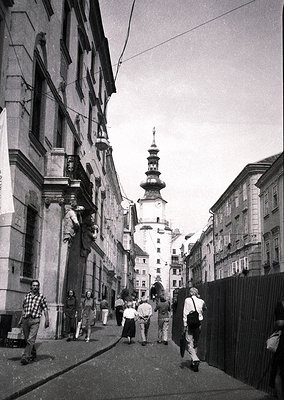 Narrow European street flanked by 19th-century stone buildings, leading to a Baroque-style church tower with onion dome. Crow...